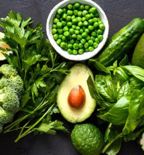 Raw healthy food clean eating vegetables: cucumber, alfalfa, zucchini, spinach, basil, green peas, dill, parsley, avocado, broccoli, lime on dark stone background, top view